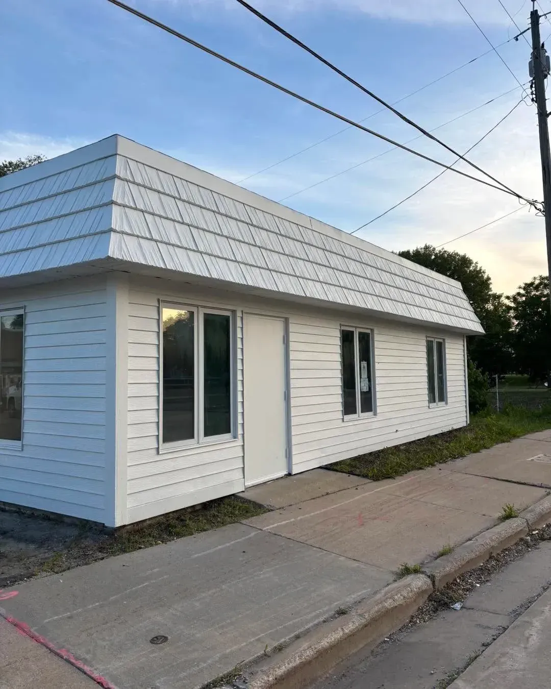 Small white house with a gray roof beside a sidewalk on a quiet street