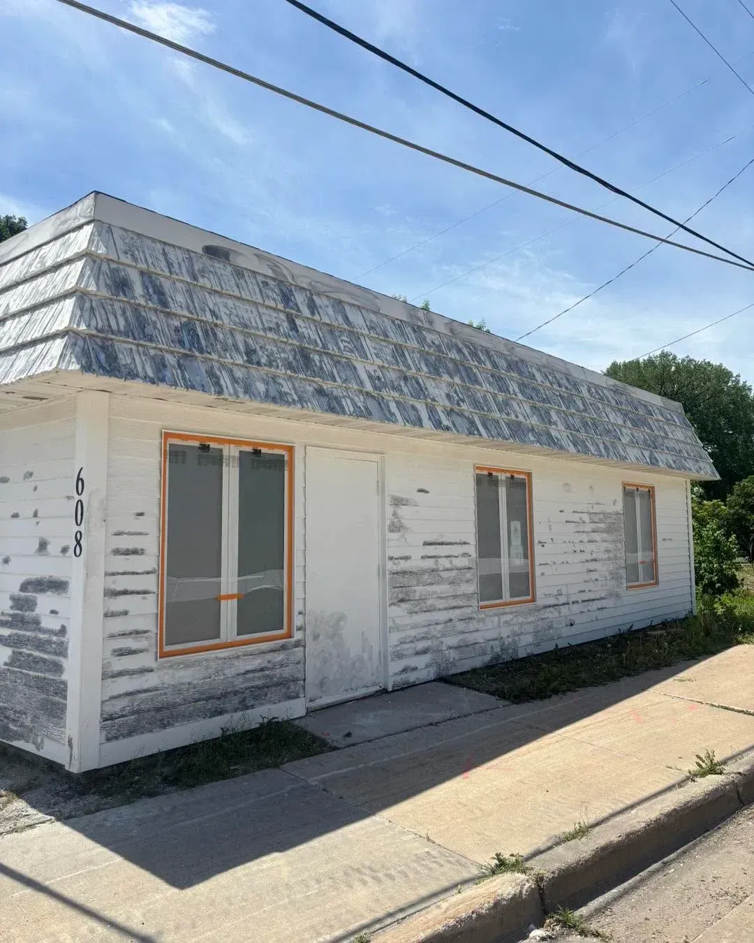 Small white house with a weathered roof, two windows, and a sidewalk in front.