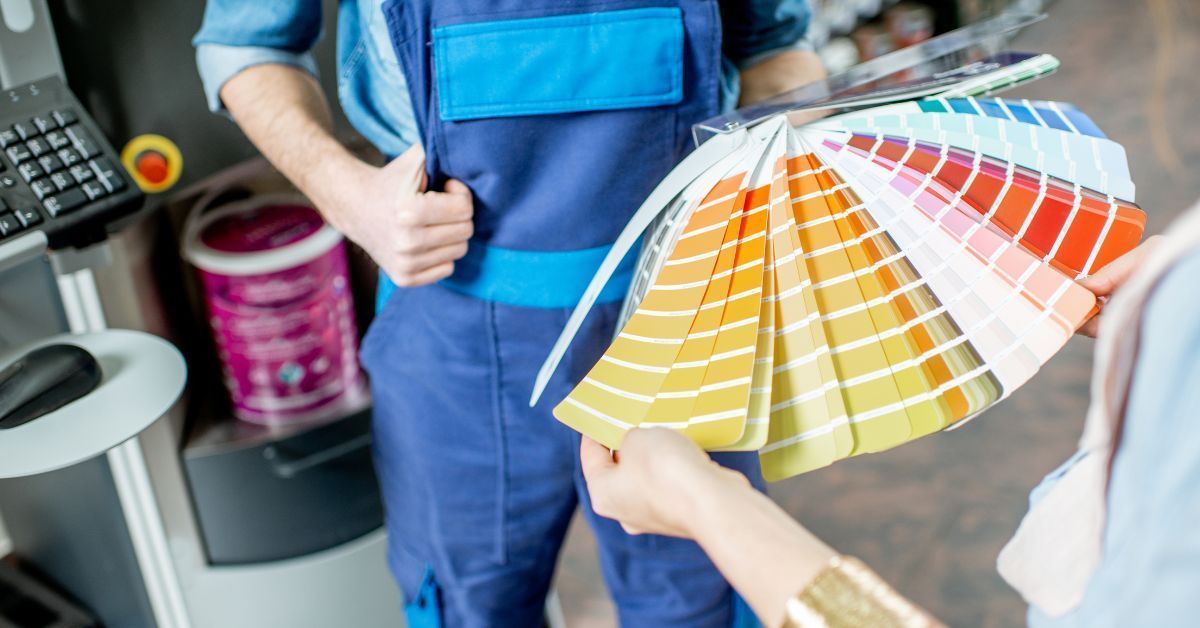 Person in blue overalls showing paint color samples to a person. Interior, paint machine visible.